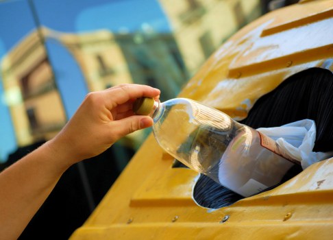 A person's hand, wearing a beige glove, is seen placing a clear plastic bottle into a yellow waste collection container mounted on a vehicle. The bottle appears empty, with some residual liquid visible inside, and has a green cap. The container has a small circular opening designed for disposing of bottles, with part of a paper bag lodged beside the opening. The vehicle's surface is painted yellow and has a smooth, glossy finish, reflecting sunlight. In the background, there are blurred building structures and windows, indicating an outdoor urban environment, possibly on a street in Blackwall or nearby areas. The scene captures a moment of rubbish disposal, aligning with commercial waste removal activities conducted by services like Commercial Waste Blackwall, particularly focused on plastic waste collection as part of business waste management.