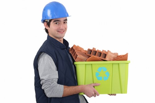 A young male waste collection worker wearing a blue safety helmet and a dark blue vest over a grey long-sleeve shirt is standing against a plain white background. He is smiling slightly and holding a bright green recycling bin with a blue recycling symbol on the side, filled with red clay bricks. The bricks are stacked neatly but are visibly rough-textured and rectangular, with some small gaps between them. The bin's vibrant color contrasts with the neutral tones of the worker's clothing and the reddish-brown bricks. His left arm supports the base of the bin, while his right hand points at the bin’s side, emphasizing its recyclable content. The scene appears staged for promotional or informational purposes related to rubbish removal and recycling services, with a focus on responsible waste disposal practices relevant to the Blackwall area of London. The clean, well-lit image highlights the practical aspects of rubbish collection, suitable for a commercial waste management website.