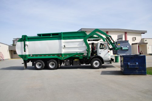 A large commercial rubbish collection truck operated by Commercial Waste Blackwall is parked on a paved street in front of a modern residential building. The truck features a predominantly white cargo area with green accents along its edges and frame, and is equipped with a hydraulic arm mechanism extending towards a large blue dumpster positioned on the right side of the image. The hydraulic arm appears to be in the process of lifting or preparing to empty the dumpster, which is situated on the asphalt surface of the driveway or street. The truck's cab is grey, and the vehicle has six wheels visible—two in the front and four at the rear—matching the purpose of waste collection services in the local area. The surrounding environment includes a contemporary residential building with white and beige exterior walls, a flat roof, and minimal architectural details, indicating a suburban or urban setting. The lighting suggests an overcast sky, and there are no other vehicles or pedestrians visible, emphasizing the truck’s active role in rubbish removal services near Blackwall or the surrounding postcode area.