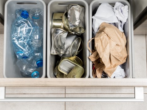 The image shows three grey plastic bin liners filled with different types of waste, placed side by side on a white disposal unit or counter. The left liner contains empty, crumpled plastic water bottles made of clear PET plastic with blue caps. The middle liner is filled with used, empty aluminium cans, some crumpled and some upright, likely containing beverage residues. The right liner holds crumpled brown and white paper, including a crumpled brown paper bag and some white paper scraps, indicating a mix of general waste. The waste is outside a building on a tiled floor, suggesting a waste collection point or disposal area in a commercial or residential setting. The overall environment is clean and organized, with the waste neatly contained within the liners, ready for removal by a rubbish collection service such as Commercial Waste Blackwall, servicing local businesses near the postcode area.
