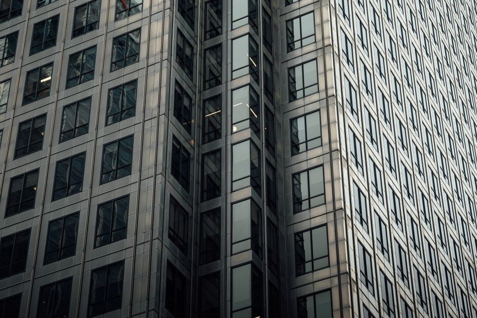 Close-up view of a modern high-rise office building with a glass and metal facade. The exterior features a grid-like pattern of reflective glass windows framed by dark metal borders, creating a sleek,