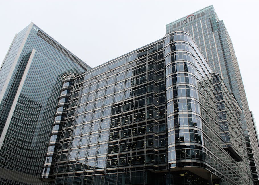 Close-up view of a modern high-rise office building with a glass and metal facade. The exterior features a grid-like pattern of reflective glass windows framed by dark metal borders, creating a sleek,