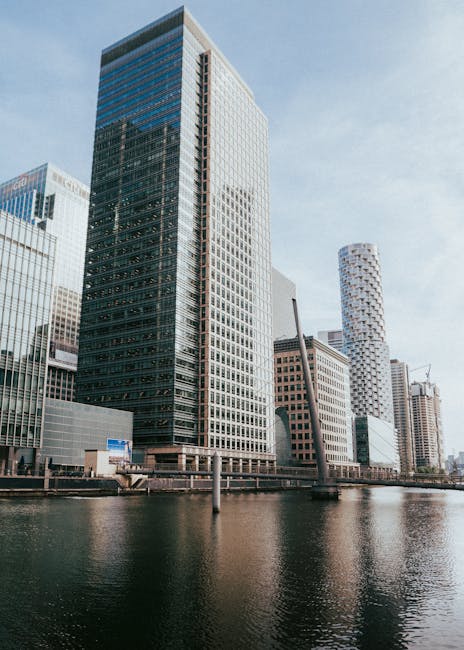 A panoramic view of a modern city skyline with numerous high-rise buildings featuring glass facades and contemporary architectural designs, situated behind a dense area of lush green trees in the fore
